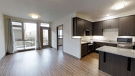 This single-floor unit features an open-concept layout that seamlessly connects the kitchen and living areas, creating a cohesive and functional living space. The kitchen is appointed with rich, dark espresso-toned cabinetry that extends to the upper walls, providing ample storage. Light-toned granite countertops offer a striking contrast against the dark cabinetry, while under-cabinet lighting enhances visibility and adds a refined touch. A stainless steel range and built-in range hood complete the kitchen's practical design. A peninsula countertop extends into the living area, offering additional workspace or casual seating. Consistent wood-look laminate flooring runs throughout, lending warmth and visual continuity to the space. The living area is bathed in natural light, courtesy of large windows and a glass-paneled exterior door that opens onto a private balcony with outdoor views. A flush-mount ceiling light fixture provides ambient illumination. An interior doorway visible from the main living space leads to an additional room, also benefiting from natural light, further enhancing the open and airy character of the unit.