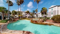 Located at 4901 Saratoga Blvd in Corpus Christi, Texas, this fitness center is a standout amenity within a lushly landscaped residential community. The facility is thoughtfully designed across a single floor and offers a comprehensive selection of exercise equipment to support a full-body workout routine. The main strength training area features cable machines, free weights, a dumbbell rack, and multiple benches, all set atop a bold blue patterned carpet that adds a polished, modern feel. Crown molding and elevated ceilings lend the space an upscale character, while recessed lighting ensures the interior is well-illuminated throughout. A dedicated cardio section includes multiple treadmills positioned beneath a wall-mounted flat-screen television, as well as a row of stationary bikes and elliptical trainers. Large windows in the cardio area frame views of the surrounding palm tree-lined grounds, filling the space with natural light. The exterior of the community showcases a resort-style pool with a natural stone fountain feature, surrounded by tall palms and manicured landscaping — creating a vibrant, tropical ambiance that complements the active lifestyle amenities within the fitness center.
