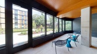Located at 404 Algoma St in Madison, Wisconsin, this single-story communal space showcases a thoughtfully designed modern aesthetic that blends natural materials with contemporary furnishings. The architecture is defined by striking angled wood-planked ceilings that lend warmth and visual depth throughout the interior, complemented by dark-toned horizontal wood wall cladding that creates a bold, cohesive look.

Expansive floor-to-ceiling windows line the perimeter, flooding the space with abundant natural light and offering unobstructed views of the surrounding landscaped grounds and mature tree canopy. The open layout flows seamlessly between distinct seating zones, including a lounge area furnished with modern upholstered sofas, mesh accent chairs, and vibrant orange and teal side tables arranged atop a colorful area rug. A built-in wood bench runs along one wall, paired with orange bar-height chairs and compact tables, creating a casual bar-style workspace or gathering area beneath pendant lighting.

A glass-walled interior room is visible through large pass-through windows, suggesting a multipurpose meeting or activity space. Exposed concrete columns punctuate the open floor plan, adding an industrial counterpoint to the warm wood tones. The polished concrete flooring ties the space together, while the overall ambiance balances relaxed comfort with energetic, community-oriented design.