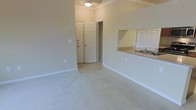 This single-story space presents a clean, open layout designed for comfortable everyday living. The main living area features light-toned wall-to-wall carpeting that contributes to a warm, neutral ambiance throughout. Crown molding adds a classic architectural touch, lining the upper perimeter of the room and lending a sense of refinement to the otherwise understated design.

A half-wall pass-through connects the living area to the kitchen, topped with granite-style countertops and equipped with a sink. A large mirror positioned above the pass-through visually expands the space, reflecting light and enhancing the sense of openness. The kitchen area transitions to tile or hard flooring, providing a practical surface distinct from the carpeted living space.

Near the entrance, a paneled front door with a deadbolt provides security, flanked by a closet that offers convenient storage directly upon entry. An additional doorway leads further into the space, suggesting access to additional rooms or a hallway. Soft ceiling lighting illuminates the area evenly. The overall palette of beige and cream tones creates a cohesive, move-in-ready environment.