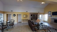 This single-floor residence welcomes visitors with a spacious, open-concept living area defined by impressively vaulted ceilings that lend a sense of grandeur and airiness to the interior. The room is finished with light-toned ceramic tile flooring that runs throughout, complementing the warm yellow walls and white upper trim to create a bright, cheerful ambiance. A ceiling fan overhead ensures year-round comfort while adding to the classic aesthetic of the space. The living area is anchored by a substantial, ornate wooden entertainment unit with open shelving, providing both display space and a sophisticated focal point. Rich leather seating and glass-topped coffee tables add a sense of refinement and ease to the room. Natural light filters in through windows along the wall, hinting at outdoor views beyond. A decorative mirror and wall art accent the space thoughtfully. A passageway leads to adjacent areas of the home, suggesting a well-connected floor plan. Overall, the space carries a warm, classic character with traditional furnishings and architectural details that make it a comfortable and welcoming environment.