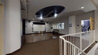 Located on W Tenth Street in Cleveland's Ohio City neighborhood, this amenity floor offers a comprehensive suite of shared facilities designed to support daily living and wellness. The centerpiece is a spacious fitness center divided into distinct workout zones. One area houses an extensive array of strength-training equipment, including cable machines, benches, and a full dumbbell rack, set against exposed brick walls and supported by bold purple-accented structural columns. A separate cardio zone features ellipticals, treadmills, and stationary bikes beneath bright linear lighting. A dedicated free-weight area with rubber flooring provides additional space for functional training. Adjacent to the gym is a multi-purpose studio room distinguished by a dramatic oval tray ceiling finished in black, housing a ceiling-mounted projector and retractable screen—suitable for fitness classes or media use. Dumbbell racks and exercise balls are neatly arranged near interior windows overlooking the fitness area. A communal lounge area features vending machines, a business workstation with multiple monitors, and framed wall art. The on-site laundry room is equipped with multiple front-load washers and stacked dryers on wood-look flooring, offering ample capacity. Throughout, the floor maintains a clean, functional aesthetic with neutral walls and practical finishes.