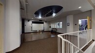 Located on W Tenth Street in Cleveland's Ohio City neighborhood, this amenity floor offers a comprehensive suite of shared facilities designed to support daily living and wellness. The centerpiece is a spacious fitness center divided into distinct workout zones. One area houses an extensive array of strength-training equipment, including cable machines, benches, and a full dumbbell rack, set against exposed brick walls and supported by bold purple-accented structural columns. A separate cardio zone features ellipticals, treadmills, and stationary bikes beneath bright linear lighting. A dedicated free-weight area with rubber flooring provides additional space for functional training. Adjacent to the gym is a multi-purpose studio room distinguished by a dramatic oval tray ceiling finished in black, housing a ceiling-mounted projector and retractable screen—suitable for fitness classes or media use. Dumbbell racks and exercise balls are neatly arranged near interior windows overlooking the fitness area. A communal lounge area features vending machines, a business workstation with multiple monitors, and framed wall art. The on-site laundry room is equipped with multiple front-load washers and stacked dryers on wood-look flooring, offering ample capacity. Throughout, the floor maintains a clean, functional aesthetic with neutral walls and practical finishes.