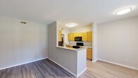 This single-floor residence is designed with a practical, open-concept layout that promotes a seamless flow between the main living areas. The entry foyer greets residents with wood-look luxury vinyl plank flooring and a front door accented by a transom window, allowing natural light to fill the space. Adjacent to the entry, the spacious living area continues the wood-look flooring and is finished in soft, neutral gray tones, creating a versatile backdrop for a variety of furniture arrangements.

The kitchen is a functional galley-style space positioned behind a peninsula-style half-wall that separates it from the living area. Warm honey-toned cabinetry lines both walls and offers generous storage, complemented by granite-look countertops and a stainless undermount sink. Black appliances, including a range, over-the-range microwave, dishwasher, and refrigerator, complete the kitchen suite. A window at the end of the kitchen brings in outdoor views and additional natural light.

The bedrooms are carpeted for comfort and include sliding-door closets with built-in wire shelving. One bedroom features a window overlooking the exterior. The bathroom presents a single-sink vanity with a large mirror, wood-tone cabinetry, and clean white finishes, connecting conveniently to the adjacent bedroom.