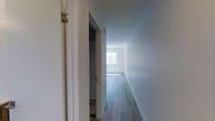 This single-floor residence offers a well-planned 1-bedroom, 1-bathroom layout making efficient use of its space. A hallway connects the main areas of the unit, finished with smooth light gray wood-look flooring that runs consistently throughout, contributing to a cohesive and modern aesthetic. The walls are painted in a clean, neutral light gray-white tone, creating a bright and airy atmosphere. White trim and door casings provide crisp contrast against the walls, adding a polished, contemporary feel to the interior. The hallway leads toward a naturally lit main living area, where a window allows daylight to filter in, enhancing the open and welcoming ambiance of the space. The clean lines and understated palette of the unit lend themselves well to a variety of furnishing styles. Overall, the unit presents a move-in-ready interior with consistent finishes, practical flow between spaces, and a neutral design that offers a versatile foundation for personalization.