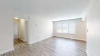This single-floor space is designed with a clean, contemporary aesthetic throughout. The open living area is finished with light-toned hardwood-style flooring and crisp white walls, creating a bright and airy atmosphere. A large window fitted with horizontal blinds allows natural light to fill the room, enhancing its spacious feel. A second, similarly styled living or dining area extends the open layout, featuring additional windows and a ceiling-mounted HVAC vent.

The galley-style kitchen is well-appointed with white upper cabinetry, warm wood-toned lower cabinets, and sleek bar-pull hardware. A classic white subway tile backsplash adds texture, while light quartz countertops and a stainless steel sink complement the stainless steel refrigerator, electric range, and dishwasher. A louvered closet door at the kitchen's end provides convenient storage.

The entryway features warm hardwood-style flooring and a louvered closet, offering practical storage upon arrival. The bedroom is carpeted in neutral tones and includes a window with blinds and a ceiling light fixture. The bathroom is finished with wood-look flooring, a light wood vanity with a white countertop, and a tub-shower combination with a curved curtain rod.
