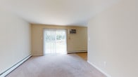 This single-floor unit offers a practical and comfortable layout with a neutral color palette of warm beige tones throughout. The living area is generously sized, featuring wall-to-wall carpeting and a full-length sliding glass door dressed with vertical blinds, flooding the space with natural light and providing views of the surrounding greenery. Baseboard heating and wall-mounted air conditioning units ensure year-round climate control.

A hallway entry welcomes residents through a paneled front door with a glass transom, transitioning from hardwood-style flooring into the main carpeted living spaces. The kitchen is well-appointed with warm honey-toned wood cabinetry extending to the ceiling, granite-style countertops, a double-basin sink, and white appliances including a refrigerator and electric range. Hardwood-style flooring runs through the kitchen, which opens toward a laundry area.

The bedrooms are carpeted and well-proportioned, each offering natural light through windows. One bedroom features dual closets, including a walk-in and a standard closet, providing excellent storage capacity. The bathroom is clean and functional, finished with light tile flooring, a white vanity with cabinet storage, and a shower-tub combination. Overall, the space presents a straightforward, move-in-ready layout.