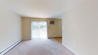 This single-floor unit offers a practical and comfortable layout with a neutral color palette of warm beige tones throughout. The living area is generously sized, featuring wall-to-wall carpeting and a full-length sliding glass door dressed with vertical blinds, flooding the space with natural light and providing views of the surrounding greenery. Baseboard heating and wall-mounted air conditioning units ensure year-round climate control.

A hallway entry welcomes residents through a paneled front door with a glass transom, transitioning from hardwood-style flooring into the main carpeted living spaces. The kitchen is well-appointed with warm honey-toned wood cabinetry extending to the ceiling, granite-style countertops, a double-basin sink, and white appliances including a refrigerator and electric range. Hardwood-style flooring runs through the kitchen, which opens toward a laundry area.

The bedrooms are carpeted and well-proportioned, each offering natural light through windows. One bedroom features dual closets, including a walk-in and a standard closet, providing excellent storage capacity. The bathroom is clean and functional, finished with light tile flooring, a white vanity with cabinet storage, and a shower-tub combination. Overall, the space presents a straightforward, move-in-ready layout.
