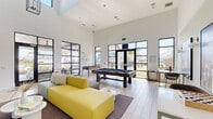 This contemporary lounge is a single-floor amenity space designed for recreation and social gathering. The room's most striking architectural feature is its dramatically high ceiling, which amplifies the sense of openness and gives the interior an airy, expansive feel. Crisp white walls serve as a clean backdrop, complemented by large black-framed glass windows and doors that wrap around the perimeter, flooding the space with natural light and providing unobstructed views of the surrounding outdoor environment.

Light-toned tile and wood-style flooring runs throughout, reinforcing the bright, modern aesthetic. The lounge is thoughtfully appointed with a full-size pool table positioned centrally, serving as a natural focal point, while a foosball table occupies a corner for additional entertainment. Modular seating in bold hues of yellow, mint, and magenta is arranged to encourage casual conversation and relaxation.

Wall-mounted flat-screen televisions are paired with a sleek floating media console, offering a dedicated viewing area. Oversized black-and-white photographic artwork and a colorful neon wall installation add artistic character to the space. High-top tables and bar-height seating along the perimeter provide versatile spots for casual use. Overall, the lounge balances energetic design with functional, well-scaled amenities.