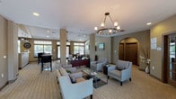 This community room is designed to serve as a versatile, welcoming gathering space for residents. The main lounge area is anchored by a sophisticated circular chandelier and features elegant, neutral-toned seating arrangements, including plush sofas and accent chairs grouped around glass-top coffee tables — ideal for socializing or relaxing. Structural columns and a distinctive arched doorway add architectural character to the space, while recessed lighting and large transom windows allow natural light to fill the room throughout the day. French doors at the rear open to outdoor views, further enhancing the open, airy atmosphere. A dedicated reception or leasing desk area is integrated seamlessly into the lounge, providing a functional workspace within the communal setting. A separate entertainment lounge offers a wall-mounted flat-screen TV, comfortable seating, a ceiling fan, and additional French doors with garden views — perfect for casual gatherings. The fully equipped kitchen features dark cabinetry, light stone countertops, black appliances including a range and refrigerator, a double sink, tile flooring, and ample counter space, making it well-suited for events and catered functions. Together, these distinct zones create a cohesive, multi-purpose community environment that balances comfort, functionality, and style.