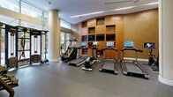 Located at 2101 N Westmoreland St in Arlington, Virginia, this fitness center offers a thoughtfully designed exercise space across a single floor. The layout is defined by a graceful curved accent wall clad in warm wood paneling and shimmering gold mosaic tile, creating a striking visual centerpiece that anchors the cardio zone. Floor-to-ceiling windows flood the space with natural light and provide views of the surrounding greenery, while cylindrical columns and recessed linear lighting lend a modern, polished aesthetic.

The cardio area is well-equipped with multiple treadmills, ellipticals, a stair climber, and Peloton cycling bikes, many featuring integrated touchscreens and backed by built-in shelving units with mounted flat-screen televisions. The strength training section offers a comprehensive selection of free weights, a dumbbell rack, weight benches, cable machines, and a multi-station strength system. Full-length mirrors along one wall visually expand the space and support form monitoring during workouts.

Durable rubber flooring covers the entire exercise area, providing comfort and safety underfoot. A bold orange accent wall near the entrance adds an energetic pop of color, complemented by organized storage for yoga mats and medicine balls. The overall ambiance balances functionality with upscale design, making it a well-rounded facility for a variety of fitness routines.
