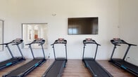 Located at 12832 White Willow Dr in Plainfield, IL, this single-floor fitness facility is thoughtfully designed to support a well-rounded exercise routine. The space is divided into distinct workout zones, each finished with durable wood-look flooring and crisp white walls that create a clean, motivating atmosphere throughout.

The cardio area is equipped with multiple treadmills arranged neatly along one wall, each featuring an individual digital display console. A large wall-mounted flat-screen television is positioned prominently above the treadmills, allowing users to stay entertained during their workouts. High ceilings in this section enhance the sense of openness, while interior glass panels and doorways connect to adjacent areas, maintaining visual flow throughout the facility.

A dedicated cycling zone houses several stationary spin bikes arranged to face a generous bank of tall, multi-pane windows. These windows flood the room with natural light and offer views of the surrounding neighborhood streetscape, creating an energizing environment for indoor cycling sessions.

An additional open exercise room provides a flexible, mat-friendly space for stretching, yoga, or floor exercises. This room benefits from a large triple-pane window framing tranquil views of an open green landscape, contributing a calm and airy quality to the space. Together, the zones offer a complete and well-appointed community fitness experience.