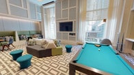 This thoughtfully designed amenity lounge combines sophisticated aesthetics with recreational functionality across a single open floor. The space immediately impresses with its dramatically tall ceilings, adorned with a structured paneled wall treatment in soft grey and white tones that adds architectural depth and elegance throughout. Large floor-to-ceiling windows dressed in sheer white curtains flood the interior with abundant natural light while framing views of the surrounding cityscape.

The lounge is anchored by a regulation billiards table with a striking teal felt surface, positioned beneath sleek pendant lighting for optimal play. Multiple distinct seating zones encourage both social gatherings and relaxed lounging, featuring a mix of modular sofas in grey and chartreuse green, teal accent ottomans, and plaid throw pillows that add playful pops of color. A built-in banquette finished in sage green upholstery offers an intimate nook alongside a round wood bistro table and contemporary chairs.

Two wall-mounted flat-screen televisions are thoughtfully positioned for viewing from multiple areas of the room. The boldly patterned geometric carpet in grey and cream ties the various seating zones together cohesively. A separate kitchenette area features a clean white quartz countertop with an undermount stainless steel sink, providing convenient refreshment support for the lounge.