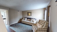 This two-story home offers a practical and comfortable layout spread across two levels, connected by a staircase visible in the floor plan. The living room on the main level features dark hardwood-style flooring, neutral beige walls, and a textured ceiling, creating a warm and relaxed atmosphere. A decorative mirror anchors the main wall, complementing the casual seating arrangement.

Adjacent to the living area, the dining space accommodates a rustic industrial-style table with matching stools, positioned on a textured area rug. The kitchen is efficiently designed with gray cabinetry, gold hardware, granite-patterned countertops, a stainless steel sink, and white appliances including a range and refrigerator, all set over matching hardwood-style flooring.

Upstairs, the bedrooms feature plush carpeted floors and neutral-toned walls. One bedroom presents a dark wood furniture set, while another is outfitted with crisp white furniture and soft, light-toned décor. A third bedroom offers a window that brings in natural light. The bathroom includes a full tub-shower combination with clean white subway tile surround and chrome fixtures, maintaining a simple and functional aesthetic throughout.