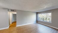 This single-floor residence offers a clean, functional layout with consistent finishes throughout. The living area is generously proportioned, featuring light wood-style flooring, neutral gray walls, crisp white baseboards, and a textured ceiling. A large picture window floods the space with natural light and frames views of lush, mature landscaping outside. The open layout connects the living area to the kitchen, which is separated by a peninsula counter and equipped with light wood cabinetry, white appliances including a gas range, microwave, and refrigerator, a stainless steel sink, and light-toned countertops. A ceiling fan in the adjacent dining or secondary living area adds both comfort and airflow. The bedroom is a comfortable retreat, offering a wide mirrored sliding-door closet for ample storage and a horizontal clerestory-style window that brings in natural light while maintaining privacy, with views of the well-maintained greenery beyond. The full bathroom features a white vanity with a speckled countertop, a bathtub with shower, and clean, simple finishes. Consistent light wood flooring flows through the majority of the space, tying each room together with a warm, cohesive aesthetic.