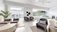 This single-level amenity space is designed with a clean, contemporary aesthetic, characterized by an all-white color palette, polished concrete flooring, and high ceilings that lend an open, airy atmosphere throughout. The layout flows seamlessly between distinct zones, each thoughtfully furnished to encourage relaxation and social interaction.

The poolside lounge area opens directly toward the outdoor pool, separated by metal railing fencing that allows unobstructed views of the water. Ceiling fans overhead provide comfort in this semi-open setting, which is furnished with woven rattan seating, a sculptural mosaic coffee table, and patterned area rugs. Decorative black circular wall art adds a bold focal point to the otherwise minimal white walls.

An interior lounge area features additional modular seating arrangements paired with round, textured ottomans, a wall-mounted flat-screen television, and lush tropical potted plants that introduce natural warmth. A water fountain station and glass double doors connect this zone to an adjacent dining or activity room visible beyond.

A dedicated games area accommodates cornhole boards and open floor space, decorated with a striking black-and-white palm tree artwork and a wooden console table, reinforcing the relaxed, resort-inspired character of the overall space.
