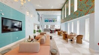 Located at 1000 East Yager Lane in Austin, TX, this single-floor amenity space is a boldly designed, artfully curated environment that blends eclectic aesthetics with functional comfort. The expansive layout is unified throughout by a striking checkered marble tile floor, which anchors the space's playful yet sophisticated design language.

The living room commands attention with soaring double-height ceilings, exposed wood beams, and walls adorned with vibrant, large-scale patterned wallcoverings in teal, orange, and gold. A teal accent wall serves as the backdrop for a flat-screen TV, complemented by a sprawling terracotta sectional sofa and a modern sputnik chandelier overhead. A seating vignette features a bold red banquette beside a marble-topped table with sculptural red chairs, while a dramatic violet marble wall panel adds a luxurious focal point. Rounded, chartreuse sculptural lounge chairs and curated artwork throughout reinforce the space's artistic character.

The office area offers a long built-in workstation lined with multiple computer terminals and cane-back task chairs beneath elegant cascading crystal chandeliers. A whimsical botanical wallpaper featuring lemons and eucalyptus leaves envelops the room, while natural light streams in through multiple windows. Decorative wooden pilasters on an adjacent wall add an architectural finishing touch to this highly functional yet design-forward workspace.