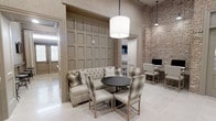 This refined single-floor space showcases a thoughtfully curated interior design that blends classic and contemporary elements. The main gathering area features a tufted banquette seating arrangement paired with a dark round table and upholstered chairs, creating an inviting social hub. A statement drum pendant light hangs overhead, casting warm, ambient light throughout the space.

Architectural interest abounds with a striking contrast between the soft, neutral-toned decorative wall paneling and a dramatic exposed brick wall, which adds warmth and texture to the environment. The light-toned tile flooring complements the overall neutral palette of creams, taupes, and grays, contributing to a cohesive and polished aesthetic.

A dedicated workstation area is nestled along the brick wall, offering a functional space equipped with a desktop computer, ideal for productivity within a comfortable setting. The ceiling height is notably generous, enhancing the sense of openness and airiness throughout. Natural light filters in through nearby doorways and openings, further brightening the interior. The overall ambiance is elegant yet approachable, blending decorative sophistication with practical, multi-use functionality.