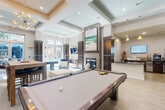 Located at 726 Hawthorne Ln in Charlotte's vibrant 28204 zip code, this single-floor amenity space offers a well-appointed and contemporary environment designed for comfort and social engagement. The layout encompasses a living room, kitchen, and hallway, all finished with a cohesive modern aesthetic.

The living room is a standout feature, boasting soaring tray ceilings with recessed lighting and an elegant sputnik chandelier. Large windows flood the space with natural light and offer views of the surrounding greenery and outdoor pool area. A sleek tile-surround fireplace anchors the room, complemented by plush blue lounge chairs, a large ottoman, and a billiards table, creating a versatile gathering space. A wall-mounted television and built-in dark wood cabinetry with mirrored accents add to the refined atmosphere.

The hallway is equally impressive, featuring wide-plank light wood flooring that transitions to large-format tile, flanked by striking dark wood columns with metallic trim details. Decorative wall medallions and recessed lighting contribute to the polished, upscale feel.

The kitchen area is outfitted with dark cabinetry, a reflective countertop, and an informational display setup complete with a wall-mounted screen — ideal for a community or hospitality setting. A distinctive driftwood horse sculpture adds an artistic touch. The overall ambiance is modern, welcoming, and thoughtfully curated.