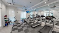 Located at 2211 Parklake Dr NE in Atlanta, Georgia, this well-appointed fitness center offers a bright and modern environment designed to support a full range of workout routines. The space is laid out on a single floor and features a clean, contemporary aesthetic with crisp white walls, recessed lighting, and ceiling fans that ensure comfortable air circulation throughout.

The main exercise area is outfitted with a robust selection of cardio equipment, including treadmills, elliptical trainers, and a recumbent bike, catering to a variety of fitness preferences. Floor-to-ceiling mirrors line one wall, visually expanding the space and allowing users to monitor their form during workouts. A dedicated section of the floor is finished with a checkered carpet tile pattern, providing cushioning underfoot for high-impact activities, while smooth hardwood-style flooring borders the perimeter for a polished finish.

Natural light flows in generously through large windows fitted with roller shades, creating an inviting and energizing atmosphere. A mounted flat-screen television is positioned for easy viewing from the cardio equipment. Additional fitness accessories, including a rack of medicine balls, are available to support strength and cross-training exercises. Large glass panel doors connect the fitness center to adjacent areas, enhancing the open and airy feel of the overall space.