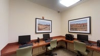 This single-floor business center is designed to provide a functional and efficient workspace for guests seeking connectivity and productivity. The room features an L-shaped wooden workstation counter that lines two walls, offering multiple individual computer setups, each equipped with a desktop monitor and keyboard. Comfortable green rolling chairs are positioned at each station, allowing for ease of movement throughout the space.

A shared multifunction printer is conveniently located at the end of the counter, accessible to all workstation users. The room is illuminated by a recessed fluorescent ceiling light, providing consistent, even lighting across the workspace. Neutral cream-colored walls contribute to a clean, distraction-free environment, while two large framed landscape prints depicting warm, earthy desert scenery add a decorative touch and visual interest to the room.

The flooring features a patterned carpet that complements the overall muted color palette. A security camera is mounted near the ceiling, reflecting attention to guest safety. The space is compact yet thoughtfully arranged to maximize functionality within its footprint.