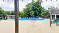 Located at 501 Highway 54 in Carrboro, North Carolina, this outdoor pool facility offers a well-appointed recreational amenity set within a naturally lush, tree-lined environment. The centerpiece of the space is a generously sized swimming pool with crystal-clear blue water, featuring a designated lap lane for fitness swimming. The pool is framed by a wide concrete deck that provides ample space for lounging and circulation.

Surrounding the pool deck is an extensive collection of teal lounge chairs and seating clusters, complemented by vibrant orange patio umbrellas that provide shade throughout the day. The layout accommodates a large number of guests comfortably, making it well-suited for both relaxed leisure and active recreation.

A brick-accented clubhouse building anchors one side of the pool area, featuring large windows and glass doors that connect the interior to the outdoor space. An additional covered pergola or shade structure is visible nearby, offering a more sheltered gathering area. The entire pool area is enclosed by a black metal fence, ensuring privacy and security.

Towering mature trees border the perimeter, creating a natural green backdrop that adds to the tranquil ambiance of the space. The combination of well-maintained facilities, generous seating, and a scenic natural setting makes this an inviting and functional outdoor amenity.