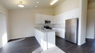 This single-floor space showcases a clean, contemporary kitchen designed for both functionality and style. The kitchen is anchored by a central island that doubles as a prep station, complete with a built-in sink and light granite-style countertops that contrast beautifully against the dark hardwood flooring below. White flat-panel cabinetry lines the walls from counter to ceiling, providing generous storage while contributing to the space's bright, airy aesthetic. Stainless steel appliances, including a range with cooktop, an over-the-range microwave, and a full-size refrigerator, lend a polished, modern finish. Recessed lighting in the ceiling is complemented by a pendant light fixture near the island, ensuring the space is well-lit throughout. The neutral warm-toned walls enhance the sense of openness, while natural light streams in, further illuminating the kitchen. The dark hardwood floors ground the space and add a rich contrast to the predominantly white elements. The overall design balances practicality with modern style, making it an inviting and efficient space.