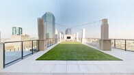 Located atop The Yard at 969 Commerce St in downtown Fort Worth, this rooftop terrace is a sophisticated open-air amenity space that combines lush greenery with modern design. A generously sized artificial lawn anchors the center of the terrace, providing a vibrant focal point surrounded by wide-format stone tile walkways and warm wood deck sections. String lights span the length of the space overhead, adding an inviting ambiance suited for both daytime lounging and evening gatherings. A built-in outdoor entertainment wall featuring a large flat-screen TV serves as a social hub, flanked by neutral-toned lounge chairs and seating arrangements. Multiple distinct seating zones are distributed across the terrace, including plush modular sofas with a central fire table on the wood deck area, as well as chaise lounges and accent chairs positioned throughout. Glass panel railings along the perimeter ensure unobstructed, panoramic views of the Fort Worth skyline, with glittering high-rise towers and the broader cityscape visible in every direction. The building's limestone-clad facade rises alongside the terrace, reinforcing an upscale architectural character. The overall atmosphere is bright, open, and resort-inspired, ideal for relaxation and social events above the heart of the city.