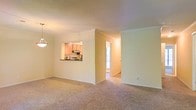 This single-floor residence presents a clean, open layout designed for comfortable everyday living. The main living area is finished with wall-to-wall carpeting in a neutral tone, creating a warm and cohesive feel throughout the space. Crown molding runs along the ceiling, adding a subtle architectural detail that elevates the overall finish of the interior.

The kitchen is accessible via a pass-through window that opens directly into the living area, allowing for easy communication between spaces and maintaining an open, connected atmosphere. The kitchen features light wood cabinetry and under-cabinet task lighting, providing both functionality and a bright working environment.

Adjacent to the main living space, a hallway leads to additional rooms, one of which benefits from natural light filtering through a window with blinds, contributing to a bright and airy ambiance. The walls are painted in a soft, warm neutral tone that complements the carpeting and cabinetry, creating a harmonious and inviting interior palette throughout.

Overall, the space is thoughtfully laid out on a single level, making it highly accessible and easy to navigate. The combination of practical finishes, open sightlines, and natural light makes this a welcoming and functional home.