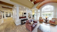Located at 5617 111 St E in Puyallup, Washington, this single-floor community clubhouse offers an impressive blend of architectural detail and functional amenity spaces. The main gathering area is defined by soaring vaulted ceilings adorned with rich, exposed wood beam accents and a striking arched tongue-and-groove wood ceiling inset, creating a warm and sophisticated atmosphere. Tall white columns delineate the open-plan layout, guiding residents through interconnected lounge areas finished with hardwood-style flooring and neutral carpeting.

Multiple seating areas are furnished with rich leather sofas and chairs, providing comfortable spaces for socializing. A focal-point fireplace with a tile surround, classic white mantel, and flanking built-in shelving anchors one of the primary lounge areas. Large, multi-pane windows and floor-to-ceiling glass panels throughout the space invite abundant natural light and offer views of the outdoor patio and landscaped surroundings.

A well-appointed kitchen features granite countertops, warm wood cabinetry, a tile backsplash, and stainless steel appliances, ideal for community gatherings. A bright dining nook with expansive windows overlooks the outdoor patio area. Additional amenities include a business center with dual computer workstations and an adjoining fitness center equipped with cardio and strength-training equipment, visible through a large interior window.