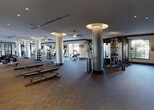 Located at 6600 Main St in Miami Lakes, FL, this well-appointed fitness center offers a comprehensive, single-floor layout designed to accommodate a wide variety of workout routines. The space is characterized by its generous open floor plan, supported by distinctive cylindrical columns with illuminated disc accents that add an architectural flair to the interior.

The cardio zone is equipped with a row of treadmills and additional cardio machines, each positioned beneath large wall-mounted televisions for an engaging workout experience. Ceiling fans throughout the space promote comfortable air circulation during intense sessions.

A dedicated free weights area features an extensive dumbbell rack alongside flat benches and floor space for stretching, all enhanced by full-length mirrors that amplify the sense of space. A separate strength training section houses a multi-station cable and resistance machine setup, ideal for targeted muscle group training.

An additional room, visible through large glass panel partitions, provides space for cycling machines and supplemental equipment. Large windows along the perimeter walls invite abundant natural light and offer views of the lush surrounding greenery, creating an energizing atmosphere. The durable carpeted flooring and neutral color palette contribute to a clean, professional ambiance throughout.