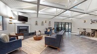 Located at 46 Avonwood Road in Avon, Connecticut, this thoughtfully designed single-floor community clubhouse offers a diverse range of amenities within an architecturally striking setting. The centerpiece of the space is a expansive main lounge area distinguished by soaring vaulted ceilings with exposed white-painted beam trusses and skylights that flood the interior with natural light. Wide-plank hardwood-style flooring runs throughout, lending warmth and continuity. A tile-surround fireplace anchors one end of the lounge, complemented by comfortable seating arrangements and a wall-mounted television. Multiple dining and high-top table groupings provide flexible gathering options. Large glass doors connect the lounge directly to a well-equipped fitness center, maintaining an open and airy feel. A dedicated billiards room features a full-size pool table beneath an elegant chandelier, bold accent walls, and built-in cue storage. Adjacent to it, a game nook with high-top tables and ample natural light through slatted windows offers space for casual recreation. The facility also includes a compact, fully equipped kitchen with white shaker cabinetry, dark granite countertops, and stainless steel appliances. Transition areas feature clean stairway landings with handrails and framed artwork, ensuring a polished, cohesive aesthetic throughout.