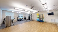 Located at 3640 Harmony Commons Drive in Spring, TX, this fitness center offers a thoughtfully organized, single-floor layout designed to support a full range of workout routines. The space is divided into two distinct functional zones: a carpeted exercise area and an open hardwood-style floor section, providing versatility for both equipment-based and free-movement training.

The cardio and strength zone is equipped with multiple treadmills, a stationary upright bike, a multi-function cable machine, and a dumbbell rack, giving users a comprehensive selection of resistance and aerobic options. A dedicated leg press machine is also present near the entrance area. The open floor zone offers ample room for stretching, yoga, or group exercise, complemented by a well-stocked rack of medicine balls, resistance bands, and stability balls in various sizes.

The room is brightly lit with overhead fluorescent tube lighting and features multiple ceiling fans to maintain airflow throughout. Natural light enters through several windows fitted with blinds, while a wall-mounted flat-screen television adds a modern touch. Double glass doors provide convenient access to an adjacent outdoor area. The neutral-toned walls and clean finishes create a welcoming, functional environment suitable for a broad range of fitness activities.