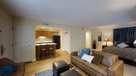 This single-floor residence offers a well-designed open-concept layout that seamlessly connects the main living areas. The living room is a comfortable and inviting space, featuring warm hardwood-style flooring, recessed lighting, and neutral wall tones that create a bright, clean ambiance. A plush sofa with accent pillows anchors the seating area, complemented by additional ottomans and soft area rugs that add texture and warmth.

The kitchen is efficiently designed and open to the living space, featuring warm wood cabinetry, a black refrigerator, and a peninsula countertop with bar-height seating for two — ideal for casual dining or entertaining. Recessed ceiling lights illuminate the workspace, and a window above the sink allows for natural light.

Crown molding runs along the ceiling throughout the main living area, adding a refined architectural detail. HVAC vents are thoughtfully integrated into the ceiling, ensuring year-round comfort. Doorways visible from the main space suggest additional rooms, including what appears to be a bedroom, offering a practical and functional floor plan. Overall, the space presents a clean, cohesive aesthetic that balances everyday functionality with comfortable living.