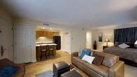 This single-floor residence offers a well-designed open-concept layout that seamlessly connects the main living areas. The living room is a comfortable and inviting space, featuring warm hardwood-style flooring, recessed lighting, and neutral wall tones that create a bright, clean ambiance. A plush sofa with accent pillows anchors the seating area, complemented by additional ottomans and soft area rugs that add texture and warmth.

The kitchen is efficiently designed and open to the living space, featuring warm wood cabinetry, a black refrigerator, and a peninsula countertop with bar-height seating for two — ideal for casual dining or entertaining. Recessed ceiling lights illuminate the workspace, and a window above the sink allows for natural light.

Crown molding runs along the ceiling throughout the main living area, adding a refined architectural detail. HVAC vents are thoughtfully integrated into the ceiling, ensuring year-round comfort. Doorways visible from the main space suggest additional rooms, including what appears to be a bedroom, offering a practical and functional floor plan. Overall, the space presents a clean, cohesive aesthetic that balances everyday functionality with comfortable living.