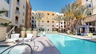 This outdoor amenity space presents a well-appointed resort-style pool area designed for relaxation and social engagement. The centerpiece is a generously sized swimming pool with clear, blue water, framed by a broad, paved pool deck finished in neutral tones that complement the surrounding architecture. Tall, mature palm trees are positioned throughout the courtyard, providing shade and a distinctly warm-climate ambiance under open skies.

The pool deck is furnished with contemporary lounge chairs and chaise lounges, offering ample space for sunbathing. Several shaded cabanas with draped curtains provide private seating areas equipped with wicker lounge chairs and glass-topped tables, creating comfortable spots for gathering. Additional gazebo-style shade structures are dispersed around the perimeter, each offering seating beneath canopied tops.

Lush desert landscaping lines the borders of the space, featuring agave, succulents, ornamental grasses, and tropical plants such as bird-of-paradise, adding texture and greenery. The surrounding multi-story building features warm earth-tone façades with stone accent walls and large windows, enclosing the courtyard and creating a sense of privacy. The overall atmosphere is modern, polished, and evocative of an upscale resort environment.