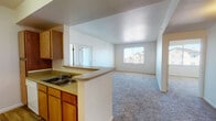 This single-floor residential unit offers a functional and well-organized layout with an open, connected flow between its main living spaces. The kitchen is a standout feature, equipped with warm wood-toned cabinetry, a double-basin sink, a full suite of white appliances including a range and refrigerator, and durable wood-look flooring. A peninsula counter extends into the adjoining space, creating a natural divide between the kitchen and the carpeted living area. The living room is bright and welcoming, benefiting from multiple windows that invite natural light and offer views of the surrounding community. A dedicated entry door with a window panel provides additional light and access to the exterior. Multiple bedrooms are spread throughout the unit, each featuring plush carpeting, neutral walls, and windows that allow ample daylight. The rooms offer comfortable, versatile spaces suitable for a variety of uses. The bathroom is cleanly appointed with a tub-shower combination accented by white tile surround, grab bars, a single-sink vanity with wood cabinetry, and wood-look flooring. Central HVAC vents are present throughout, ensuring year-round climate comfort. The overall aesthetic is clean, neutral, and move-in ready.