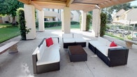 This covered outdoor lounge space is defined by a striking pergola structure supported by substantial stucco columns, creating a shaded retreat that seamlessly connects interior comfort with the surrounding landscape. The slatted overhead beams allow filtered natural light to play across the concrete floor throughout the day, lending the space a warm, inviting ambiance.

The lounge area is furnished with a generously sized modular wicker sectional seating arrangement, complete with plush white cushions and accented with bold red throw pillows. A dark wood-topped coffee table anchors the arrangement, making the space well-suited for casual conversation or relaxation. Stone benches along the perimeter of the pergola provide additional seating and contribute to the architectural character of the area.

Climbing vines and mature greenery weave around the columns, softening the structure and integrating it naturally into the well-maintained lawn and landscaped surroundings. Views of manicured hedgerows and adjacent buildings are visible through the open colonnades, reinforcing the sense of being part of a larger, thoughtfully designed outdoor environment. Overall, this space offers a functional and aesthetically cohesive outdoor gathering area with a strong sense of place and comfort.