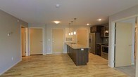 This single-floor space is designed around an open-concept layout that seamlessly integrates the kitchen and living areas. The kitchen serves as the focal point, anchored by a large central island finished with granite countertops and a built-in sink. Dark cabinetry on the island contrasts attractively with the light stone surface, while a trio of pendant lights hang above, providing both task lighting and a decorative element.

Along the kitchen's perimeter, dark wood upper and lower cabinetry is paired with stainless steel appliances, including a French door refrigerator, a range with an oven, and an over-the-range microwave. Recessed lighting complements the overall illumination throughout the space.

Light-toned hardwood flooring extends across the entire unit, lending warmth and a sense of continuity to the open floor plan. The neutral wall colors contribute to a clean, modern aesthetic. Multiple doors visible from the main living area suggest access to additional rooms, storage, or building corridors. Overall, the space offers a functional and contemporary layout suited for everyday living.