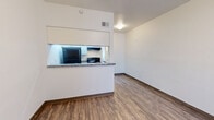 This single-story unit offers a clean, functional layout designed for comfortable everyday living. The open-concept living and dining area is finished with durable wood-look flooring and crisp white walls, creating a bright and welcoming atmosphere. A pass-through kitchen anchors the living space, featuring granite-style countertops, white cabinetry, a black refrigerator, an electric range with overhead ventilation, and a built-in sink — all framed by a peninsula that connects the kitchen to the main living area. Natural light enters through large windows fitted with vertical blinds, enhancing the airy feel of the space. The entry door opens directly into the main living area, providing a straightforward and accessible flow throughout. The bedroom is generously sized, outfitted with a ceiling fan with light fixture and a window with vertical blinds that allows for natural light and ventilation. A full bathroom with a shower-tub combination is conveniently accessible from the hallway. Additional utility space includes a dedicated area with built-in shelving and a water heater, offering practical storage and mechanical access. Wood-look flooring runs consistently throughout all areas, tying the space together with a cohesive, modern aesthetic.