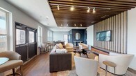 Located at 101 E Romana St in downtown Pensacola, Florida, this thoughtfully designed single-floor communal space blends modern aesthetics with functional versatility. The main lounge area is a standout, featuring rich wood-plank flooring, a striking slatted wood ceiling with integrated track lighting, and bold navy accent walls that create a sophisticated, layered atmosphere. Generous seating arrangements — including a plush sectional sofa, curved accent chairs, and a cowhide-accented coffee table — offer multiple gathering configurations suited for relaxation or social events. A large wall-mounted display screen anchors one end of the lounge, while a decorative round mirror and console table add refined detail to the accent wall. A dining nook near glass double doors provides additional seating at a bistro-style table, with the doors opening to an outdoor balcony that brings in natural light and exterior views. The kitchenette is well-appointed with stainless steel appliances, a double-basin sink, quartz countertops, dark wood cabinetry, and a striking floor-to-ceiling mosaic tile accent wall. A bar-height counter with wire-frame stools and pendant lighting completes the space, making it ideal for catered events or casual use. The overall ambiance is contemporary, polished, and inviting.