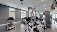 Located at 202 Onley Rd in Salisbury, Maryland, this fitness center offers a comprehensive workout environment thoughtfully organized across a generously sized single-floor space. The facility is divided into distinct training zones, each designed to support a variety of fitness routines and exercise styles.

The cardio area features a row of Matrix treadmills positioned along a gray accent wall, accompanied by elliptical trainers and stationary cycling bikes. Large windows with horizontal blinds line the walls, inviting ample natural light throughout the space and providing views of the surrounding greenery. Ceiling fans are installed throughout, contributing to comfortable airflow during workouts.

The strength training section is well-appointed with power racks loaded with Olympic weight plates, a cable machine, and a full dumbbell rack offering a wide range of weights. Wall-mounted mirrors expand the visual depth of the room and support proper form during lifts. Overhead, wall-mounted televisions provide entertainment for users.

A functional training area rounds out the facility, equipped with a heavy punching bag, medicine balls, resistance accessories, and floor matting for bodyweight and dynamic movements. The durable rubberized flooring throughout ensures safety and stability. Glass-paned doors connect the fitness floor to an adjoining corridor, and clean white and gray walls lend the space a modern, professional aesthetic.