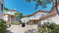 This residential complex is organized around a spacious open-air courtyard that serves as the central gathering area for its residents. The exterior features a warm stucco finish in a neutral tan tone, complemented by light teal-painted staircases and balcony railings that add a distinctive visual accent to the facade. Units are accessible from both the ground level and an upper floor, with exterior staircases providing access to second-floor balconies that overlook the courtyard below.

The shared courtyard is generously sized and finished with a smooth concrete surface, offering an open and airy communal space bathed in natural sunlight. A gas BBQ grill is positioned within the courtyard, making it a practical and social outdoor area for residents. Raised planter beds with greenery and shrubs line portions of the courtyard, adding color and softness to the hardscape environment. Mature trees provide partial shade and enhance the overall landscaping.

An arched gateway visible in the courtyard connects to an adjacent outdoor area, suggesting additional shared spaces beyond. The complex is designed with a courtyard-facing layout that fosters a sense of community while maintaining individual unit privacy.