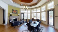 This single-floor clubhouse offers a sophisticated and inviting atmosphere designed for community gatherings and social events. The main gathering area is anchored by a striking tray ceiling with a rich, warm-toned inset and an elegant chandelier that serves as a natural focal point. Tall, multi-pane windows wrap around the room, flooding the space with abundant natural light and offering views of the surrounding exterior. Warm hardwood flooring runs throughout, complementing the space's refined yet approachable aesthetic. A classic fireplace with a white-painted surround adds a cozy, residential touch to one end of the room, accompanied by comfortable seating for intimate conversations. The central dining area features a large dark-wood table with ample seating, suitable for meetings, events, or communal dining. Accents of bold yellow-green on the walls, along with decorative artwork, lend the space a vibrant and contemporary character. Additional seating nooks near the windows provide flexible arrangements for a variety of uses. The overall ambiance is warm, light-filled, and elegantly appointed, making this clubhouse a welcoming hub for residents and guests alike.