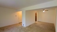 This single-floor space presents a straightforward, functional layout characterized by clean lines and neutral finishes throughout. The main living area features wall-to-wall carpeting in a light beige tone, contributing to a warm and understated ambiance. Smooth, white walls and a white ceiling keep the interior feeling bright and open.

A partial wall divider separates the living space into distinct zones, providing subtle definition without fully closing off the areas from one another. This architectural feature adds a sense of structure while maintaining an open feel between the spaces. A ceiling fan is visible in one of the adjoining areas, offering both ventilation and soft overhead lighting.

The entryway is accessible through a white door set within a recessed alcove, which also features a wall-mounted panel — likely a thermostat or intercom unit. Toward the rear of the space, a doorway leads to what appears to be a closet or additional storage area, fitted with warm wood-toned cabinetry or shelving. A floor-level vent is also present, indicating a ducted heating and cooling system. Overall, the space offers a clean, move-in ready environment with a calm, neutral palette.