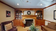 This single-floor space is defined by a striking, floor-to-ceiling custom wood built-in unit that serves as the room's central focal point. Crafted in warm honey-toned wood, the built-in surrounds a traditional fireplace with an arched surround and tile hearth, flanked symmetrically on both sides by open bookshelves stocked with an extensive collection of books. Arched display niches above each bookcase section add architectural elegance, while lower cabinet doors provide additional concealed storage.

A decorative clock is prominently displayed above the fireplace mantel, which is adorned with seasonal greenery and floral accents, adding a welcoming, lived-in character to the space. Recessed lighting is embedded throughout the ceiling, supplemented by a frosted skylight panel that contributes diffused ambient light across the room.

The flooring is covered in neutral wall-to-wall carpeting, contributing to the room's warm and comfortable atmosphere. Plush upholstered armchairs and lush potted plants complete the furnishings, creating an inviting, cozy environment well-suited for reading or relaxed conversation.