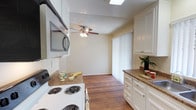 This single-floor space offers a practical and well-appointed layout with a kitchen that serves as a central feature. The kitchen is outfitted with white cabinetry providing ample storage, complemented by granite-style countertops that add a touch of warmth and texture. A built-in microwave is mounted above the electric coil range, maximizing counter space and contributing to an efficient cooking environment. A skylight above the kitchen introduces natural light, brightening the work area throughout the day.

The flooring throughout is warm-toned hardwood, lending continuity and a cohesive aesthetic as it extends from the kitchen into the adjacent living area. The living space features a ceiling fan with a light fixture, offering both airflow and ambient lighting. A sliding glass door fitted with vertical blinds allows for natural light control and likely provides access to an outdoor area.

The walls are finished in a soft, neutral tone, and the textured ceiling adds a subtle architectural detail. Overall, the space presents a clean, functional atmosphere that balances practicality with comfortable, everyday living.