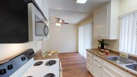 This single-floor space offers a practical and well-appointed layout with a kitchen that serves as a central feature. The kitchen is outfitted with white cabinetry providing ample storage, complemented by granite-style countertops that add a touch of warmth and texture. A built-in microwave is mounted above the electric coil range, maximizing counter space and contributing to an efficient cooking environment. A skylight above the kitchen introduces natural light, brightening the work area throughout the day.

The flooring throughout is warm-toned hardwood, lending continuity and a cohesive aesthetic as it extends from the kitchen into the adjacent living area. The living space features a ceiling fan with a light fixture, offering both airflow and ambient lighting. A sliding glass door fitted with vertical blinds allows for natural light control and likely provides access to an outdoor area.

The walls are finished in a soft, neutral tone, and the textured ceiling adds a subtle architectural detail. Overall, the space presents a clean, functional atmosphere that balances practicality with comfortable, everyday living.