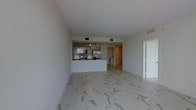 This contemporary single-floor space showcases a clean, open-plan layout that seamlessly connects the living area and kitchen, creating a sense of spaciousness and flow. The standout feature underfoot is the striking marble-effect tile flooring, with sweeping grey veining set against a crisp white background, lending an air of elegance and sophistication throughout.

The kitchen is thoughtfully designed with a peninsula-style counter that defines the cooking zone while keeping it visually connected to the main living area. White cabinetry with a high-gloss finish lines the walls, complemented by dark countertops for a bold contrast. Stainless steel appliances, including a refrigerator, range, and built-in microwave, add a polished, modern touch. Recessed track lighting above the kitchen ensures excellent task illumination.

The living area benefits from a neutral white palette across the walls and ceiling, creating a bright and versatile canvas. Recessed HVAC vents are neatly integrated into the ceiling, maintaining the clean aesthetic. A doorway visible beyond the kitchen hints at additional utility or storage space, rounding out the practical layout. Overall, the space exudes a minimalist, modern sensibility with quality finishes throughout.