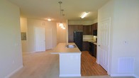 This single-floor residence offers a thoughtfully designed open-concept layout that seamlessly connects the kitchen and living areas, creating a spacious and functional environment. The kitchen serves as the focal point of the space, featuring rich dark-toned cabinetry that extends to the upper walls, providing ample storage. Black appliances, including a full-size refrigerator, range, and microwave, complement the cabinetry for a cohesive, modern aesthetic. A central kitchen island with a light countertop and integrated sink adds both workspace and casual dining potential, accented by elegant pendant lighting that hangs above. The kitchen flooring is finished in warm hardwood-style planks, which transitions into plush carpeting throughout the living area, adding comfort underfoot. The open living space is bathed in warm ambient light from flush-mount ceiling fixtures, creating an inviting atmosphere. Multiple doorways lead to additional areas of the residence, hinting at a practical and well-organized floor plan. Neutral wall tones throughout lend a clean, versatile backdrop suited to a variety of décor styles. Overall, the space balances functionality with modern design details for comfortable everyday living.
